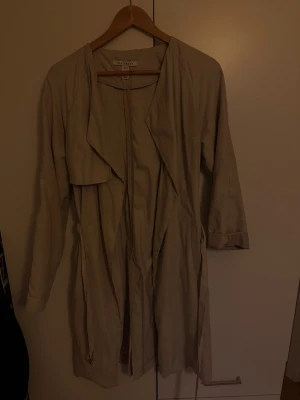 Beige trenchcoat från NLY Trend - Snygg beige trenchcoat från NLY Trend med avslappnad passform och långa ärmar. Jackan har ett vattenfallsslag och knytband i midjan för en cool siluett. Perfekt för lager-på-lager och vårens outfits.