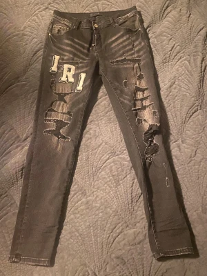 Svarta skinny jeans från Amiri med patchar - Svarta jeans från Amiri med skinny passform, slitna detaljer och stora vita bokstavspatchar på både fram- och baksida. Jeansen har dragkedja, klassiska fickor och coola broderade bokstäver som ger en unik streetkänsla.