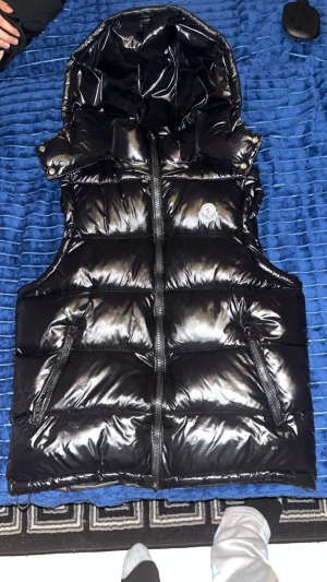 Svart pufferväst från Moncler - Svart glansig pufferväst från Moncler med huva och två dragkedjeförsedda fickor. Västen har en quiltad design och Moncler-logga på bröstet. Perfekt för att ge din outfit en lyxig vibe och hålla dig varm under kyliga dagar.