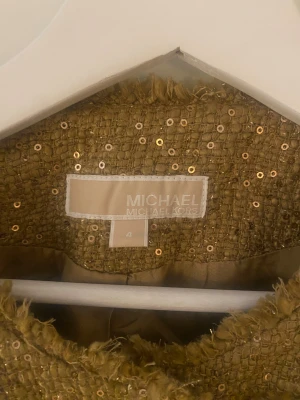 Guldig paljettkavaj Michael Kors - Snygg guldig kavaj från Michael Kors med paljetter över hela jackan. Modellen har rundad krage, råa kanter och fyra fickor framtill. Kavajen stängs med knappar och har en lyxig, glittrig look som verkligen sticker ut.