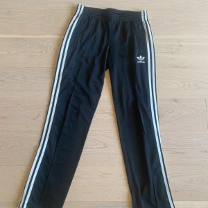 Svarta Adidas byxor med vita ränder - Svarta Adidas byxor med klassiska vita ränder längs sidorna och broderad logga på låret. Byxorna har resår i midjan, raka ben och dragkedjor vid fickorna. Perfekta för en sportig och chill stil.