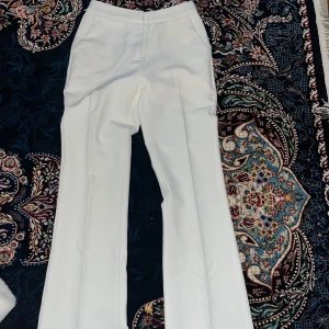 Vita kostymbyxor med flared ben  - Snygga vita kostymbyxor med flared ben och pressveck. Byxorna har diskreta fickor fram och bak, samt en klassisk midja. Perfekta för en clean och stilren look. Materialet känns mjukt och har en lätt stretch.