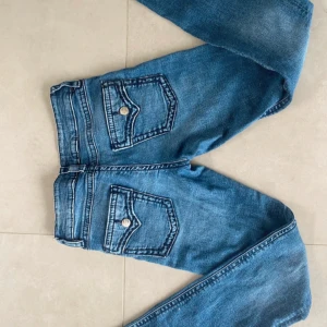 Blå jeans med fickdetaljer från Gina Tricot - Snygga blå jeans från Gina Tricot med coola fickor med lock och knapp bak. Klassisk femficksmodell med bootcut ben och normal passform. Jeansen har en mellanblå tvätt och är tillverkade i mjukt denimtyg. Perfekta för en chill och trendig look. Strl 164 men skulle klassas som en xs.