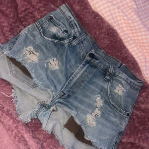 Blå slitna jeansshorts med fransig kant - Säljer ett par blå jeansshorts med hög midja och fransig kant. Shortsen har flera slitningar och hål både fram och bak, samt klassiska fickor och bälteshällor. Snygga kontrastsömmar på bakfickorna ger extra detalj. Perfekta för sommarens chill dagar.