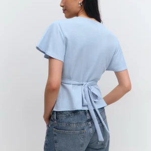 Ljusblå omlottopp från Gina Tricot - Supersöt ljusblå omlottopp från Gina Tricot med v-ringning och korta, lite fladdriga ärmar. Toppen har knytband i midjan som ger en snygg siluett. Perfekt att styla med jeans eller kjol för en chill look.