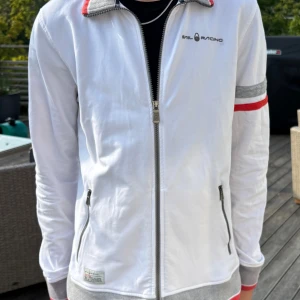 Vit Sail Racing ziptröja med detaljer - Vit långärmad ziptröja från Sail Racing med grå och röda detaljer på krage, ärmslut och midja. Tröjan har två fickor med dragkedja och en diskret logga på bröstet. Perfekt för dig som gillar sportig och stilren look. Perfekt skick. 