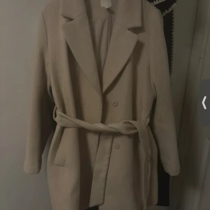 Beige kappa med bälte och knappar - Stilren beige kappa med klassisk krage, knappar framtill och ett matchande bälte i midjan. Jackan har långa ärmar och en rak passform som ger en clean look. Perfekt för dig som gillar minimalistisk stil.