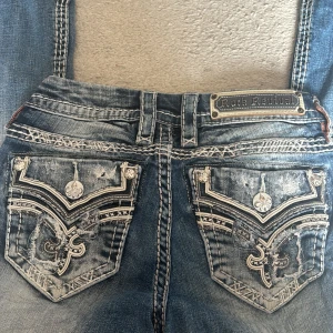 Rock Revival blå jeans med broderi - Säljer ett par blå jeans från Rock Revival med grymma detaljer. Jeansen har kontrastsömmar och broderade bakfickor med silvriga knappar och emblem. Klassisk femficksmodell i jeansmaterial med cool tvätt och snygga detaljer.