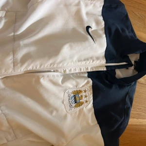 Manchester City Nike vindjacka M - Snygg vit och marinblå vindjacka från Nike med Manchester Citys klubbmärke på bröstet. Jackan har dragkedja framtill och är tillverkad i lätt polyester med meshfoder för extra komfort. Perfekt för dig som vill ha sportig stil och visa vilket lag du hejar på.