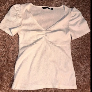 Vit v-ringad topp från Vero Moda - Superfin vit topp från Vero Moda med v-ringning och lätt rynkdetalj framtill. Toppen har korta ärmar med diskret puff och är gjord i ett ribbat, stretchigt material som sitter snyggt på kroppen. Perfekt att styla med jeans eller kjol för en fräsch look.