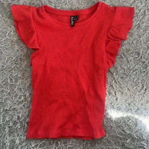 Röd ribbad topp - Superfin röd topp från Pieces med ribbad struktur och volangärmar. Modellen är figurnära och har rund halsringning. Perfekt för dig som vill ha en färgstark och söt topp med det lilla extra i ärmdetaljen.