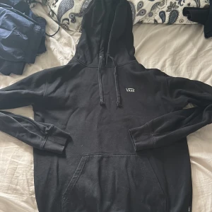 Svart hoodie från Vans med huva - Svart hoodie från Vans med klassisk logga på bröstet. Tröjan har huva med snörning och en stor magficka framtill. Långärmad och i mjukt material, perfekt för en avslappnad stil.