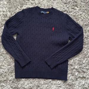 Mörkblå stickad tröja Polo Ralph Lauren - Kabelstickad mörkblå tröja Polo Ralph Lauren med klassisk rund hals och röd broderad logga på bröstet. 