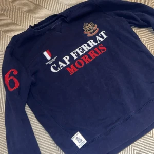 Mörkblå sweatshirt från Morris, S - Mörkblå sweatshirt från Morris med broderad text 'Cap Ferrat Morris' i vitt och rött på bröstet, samt stort rött nummer 6 på ärmen. Klassisk college-stil med ribbade muddar och detaljerade broderier. Perfekt för dig som gillar preppy look.