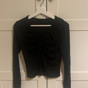 Svart långärmad topp Gina Tricot - Säljer en svart långärmad topp från Gina Tricot i storlek M. Toppen har v-ringning och snygga rynkade detaljer framtill. Materialet är stretchigt och sitter tajt mot kroppen.