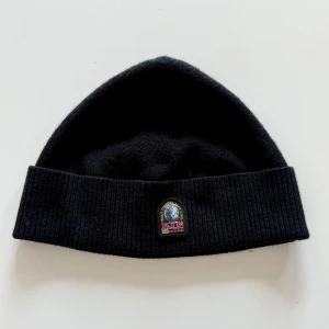 Svart mössa från Parajumpers - Svart ribbstickad mössa från Parajumpers med uppvikt kant och klassisk logopatch framtill. Perfekt för kalla dagar och enkel att matcha med olika outfits. Snygg och stilren design som funkar året runt.