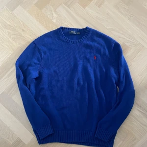 Blå stickad tröja från Polo Ralph Lauren - Klassisk blå stickad tröja från Polo Ralph Lauren med rund halsringning och röd broderad logga på bröstet. Tröjan har ribbade muddar vid ärmslut och nederkant samt lång ärm. Perfekt för dig som gillar stilren och tidlös design.