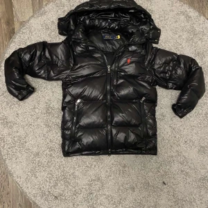 Svart pufferjacka från Polo Ralph Lauren - Säljer en svart pufferjacka från Polo Ralph Lauren med glansig finish. Jackan har huva, dragkedja framtill och två fickor med dragkedja. Klassisk quiltad design och diskret röd logga på bröstet. Perfekt för kalla dagar och riktigt snygg streetstil.