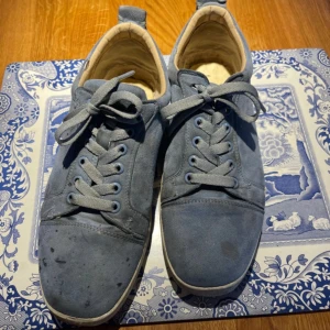 Blå mockasneakers från Christian Louboutin - Säljer ett par blå sneakers i mocka från Christian Louboutin med klassisk röd sula. Skorna har snörning, rund tå och vita kanter runt sulan. Perfekta för dig som gillar lyxiga detaljer och vill sticka ut med en ikonisk röd undersida.