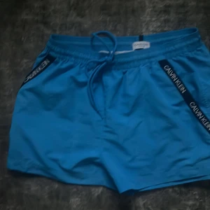 Blå badshorts från Calvin Klein - Säljer ett par blå badshorts från Calvin Klein med snörning i midjan och svarta logoband längs sidorna. Shortsen har en klassisk passform och är perfekta för stranden eller poolen. Materialet känns lätt och snabbtorkande.