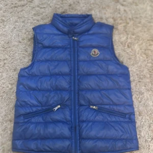 Ljusblå dunväst från Moncler - Snygg blå dunväst från Moncler med klassisk logga på bröstet. Västen har hög krage, två dragkedjefickor och quiltad design. Perfekt att slänga över en hoodie eller en stickad tröja. Stängs med dragkedja framtill. Är även i väldigt bra skick och är knappast använd. Har inget kvitto men på insidan finns en Qr kod som går att skanna för att se att den är äkta. Kan postas eller mötas upp vid intresse.