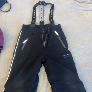 Svarta täckbyxor Polarn O. Pyret 110 - Svarta täckbyxor från Polarn O. Pyret med hängslen och reflexdetaljer längs benen och vid fickorna. Byxorna har dragkedjor, knappar i midjan och förstärkta benslut med resår och fotstrap. Perfekta för kalla dagar ute.