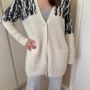 Vit och svart kofta från Noa Noa - Mysig lång kofta från Noa Noa i storlek L. Koftan är vit med svart mönster upptill och på ärmarna, har två fickor framtill och är stickad i ett mjukt material. Perfekt för lager-på-lager och chill dagar.