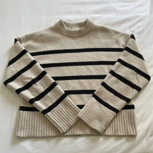 Beige och svart randig stickad tröja - Beige stickad tröja med svarta horisontella ränder och bred ribbad mudd vid ärmslut och nederkant. Tröjan har en hög, ribbad rund halsringning och lång ärm. Perfekt för dig som gillar en klassisk och avslappnad stil.