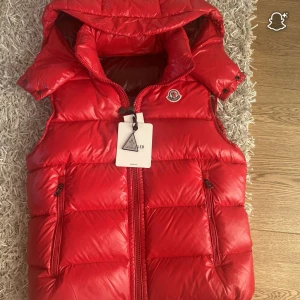 Röd dunväst från Moncler med huva - Säljer en fet röd dunväst från Moncler med avtagbar huva och två dragkedjefickor. Västen har quiltad design, Moncler-logga på bröstet och coola innerdetaljer med etiketter och serietidningstryck. Perfekt för att hålla värmen och se stylish ut.