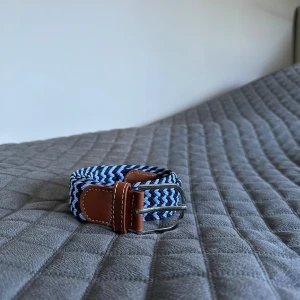 Blått flätat bälte med skinn-detaljer - Snyggt flätat bälte i olika blå nyanser med bruna skinn-detaljer och silverspänne. Bältet har ett randigt mönster och passar perfekt till jeans eller chinos. Ger en fräsch och modern look till outfiten.