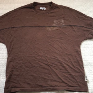 Brun Kenzo t-shirt med tigertryck - Säljer en brun t-shirt från Kenzo i storlek M. T-shirten har ett coolt tigertryck på bröstet och en markerad söm som går horisontellt över framsidan. Klassisk rund hals och korta ärmar, tillverkad i mjuk bomull. Perfekt för dig som gillar streetwear med en twist.