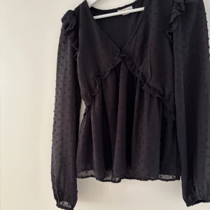 Svart blus med volang från Gina Tricot - INTRESSEKOLL! Supersöt svart blus från Gina Tricot med volangdetaljer och diskret prickig struktur. Blusen har v-ringning, långa ärmar med resår vid handlederna och en lätt transparent känsla. Perfekt för dig som gillar romantiska detaljer och flowy passform. 