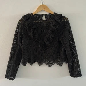 Svart spetsblus med volang från Zara - Supersnygg svart blus från Zara i spets med lång ärm och volangdetaljer framtill. Blusen har ett genombrutet mönster, rund halsringning och knapp i nacken. Perfekt för dig som gillar detaljer och vill sticka ut lite extra.
