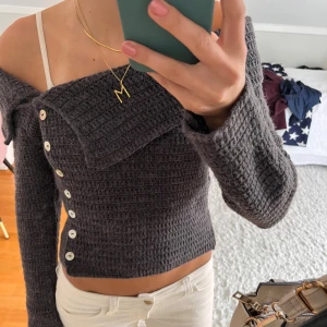 Mörkgrå offshoulder stickad tröja - Säljer en mörkgrå stickad tröja med offshoulder-design och långa ärmar. Tröjan har en croppad passform och dekorativa knappar längs ena sidan framtill. Perfekt för dig som gillar unika detaljer och en trendig look. Aldrig använd (ny utan prislapp). 