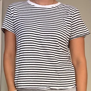 Randig vit och svart t-shirt - Säljer en klassisk t-shirt med svarta och vita horisontella ränder. T-shirten har korta ärmar och rund halsringning. Den är i en avslappnad passform och tillverkad i mjukt bomullstyg, perfekt för en chill och enkel stil.