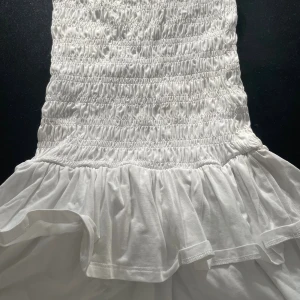 Vit volangklänning från Gina Tricot - Supersöt vit kortklänning från Gina Tricot med smockad överdel och volangig kjol. Klänningen är ärmlös och gjord i mjuk bomull, perfekt för varma dagar. Enkel och fräsch design som känns både trendig och tidlös.