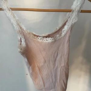 Ljusrosa ribbad linnetopp H&M Divided - Supersöt ljusrosa ribbad linnetopp från H&M Divided med vita spetsdetaljer längs axelband och ringning. Toppens material är mjukt och stretchigt, perfekt för varma dagar. Säljer då den inte kommer till användning längre!