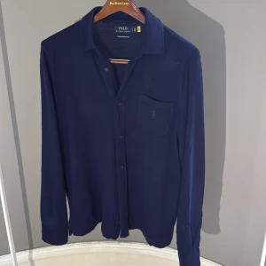 Mörkblå merinoull kofta Ralph Lauren - Snygg mörkblå kofta från Ralph Lauren i tvättbar merinoull. Klassisk krage, knäppning framtill och bröstficka med broderad logga. Perfekt för dig som gillar stilrena och bekväma plagg med premiumkänsla. Riktigt skön ger lite känsla av en overshrit i koftform nästan oanvänd 