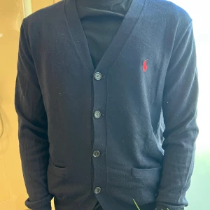 Mörkblå kofta från Polo Ralph Lauren - Snygg mörkblå kofta från Polo Ralph Lauren i 100% pima bomull. Klassisk design med knappar framtill, två fickor och röd broderad logga på bröstet. Perfekt för lager-på-lager och chill stil.