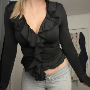 Svart omlottopp med volangdetaljer - Säljer en svart omlottopp med långa ärmar och djup v-ringning. Toppen har snygga volangdetaljer längs halsringningen och framtill, vilket ger en extra touch. Figurnära passform som framhäver midjan. Perfekt för dig som gillar trendiga och feminina plagg.