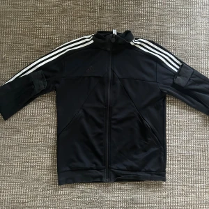 Svart Adidas track jacket med vita ränder - Svart track jacket från Adidas med klassiska vita ränder längs ärmarna och dragkedja framtill. Jackan har hög krage och två fickor på sidorna. Perfekt för en sportig och avslappnad stil.