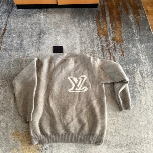Grå stickad tröja från Louis Vuitton - Snygg grå stickad tröja från Louis Vuitton med stort vitt LV-logo framtill. Tröjan har rund halsringning, ribbade muddar och är långärmad. Perfekt för dig som vill ha en stilren och exklusiv look.