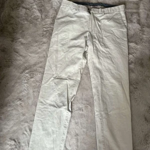 Beiga kostymbyxor från Oscar Jacobson - Snygga beiga kostymbyxor från Oscar Jacobson med raka ben och klassisk passform. Byxorna har bälteshällor, sidfickor och två bakfickor med knapp. Perfekta för en stilren och clean look. Materialet är mjukt och känns lyxigt.