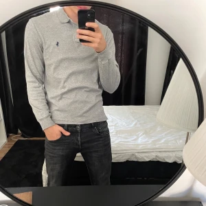 Grå långärmad pikétröja Polo Ralph Lauren - Säljer en stilren grå långärmad pikétröja från Polo Ralph Lauren. Tröjan har klassisk krage, knappar vid halsen och den ikoniska broderade loggan på bröstet. Modellen är custom slim fit och passar perfekt till jeans eller chinos.