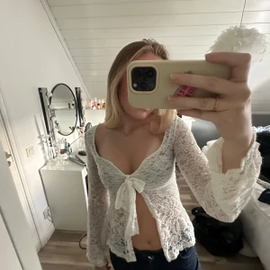 Vit spetsblus med knyt framtill - Supersöt vit blus i spets med långa ärmar och v-ringning. Blusen har ett knyt framtill och är genombruten med blommigt spetsmönster. Perfekt för dig som gillar romantisk och feminin stil. Sitter snyggt croppad och har lätt utsvängda ärmslut. Den har fått små trådar över hela toppen men går att klippa bort innan förpackning🤩storleken är xs/s men sitter som en s
