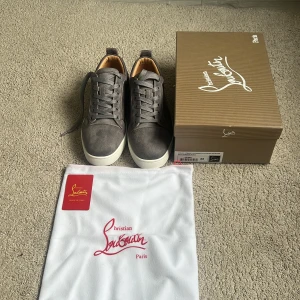 Grå vit Christian Louboutin  - HELT NYA 10/10 SKICK .Snygga gråa sneakers från Christian Louboutin.   klassisk röd sula. Skorna har snörning, rund tå och är tillverkade i mocka med vita kanter och detaljer. Perfekta för dig som vill sticka ut med exklusiv streetstyle.