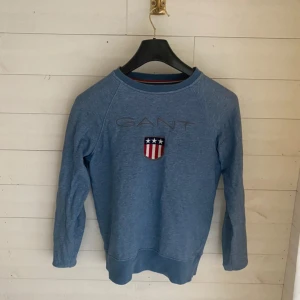 Blå sweatshirt från GANT med tryck - Blå sweatshirt från GANT med broderad logga och sköld på bröstet. Tröjan har rund halsringning och långa ärmar. Perfekt för en chill och stilren look. 