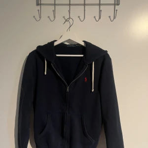 Mörkblå hoodie från Polo Ralph Lauren - Mörkblå hoodie från Polo Ralph Lauren med dragkedja framtill, vit snörning i huvan och röd broderad logga på bröstet. Klassisk passform med kängurufickor och mjukt bomullsmaterial. Perfekt för en chill och stilren look.