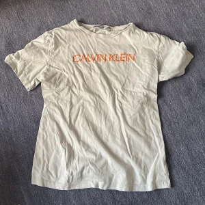 Vit t-shirt från Calvin Klein Jeans - Vit t-shirt från Calvin Klein Jeans med orange tryck på bröstet och en orange detalj med texten 'JEANS' på ryggen. Klassisk rund hals och korta ärmar. Mjuk bomullskvalitet som är skön mot huden. Perfekt för dig som gillar stilrena och coola logotoplagg.