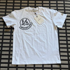 Vit Moncler T-shirt Helt Ny - Snygg vit t-shirt från Moncler med stor svart logga på bröstet och Moncler-märke på ärmen. Klassisk rund hals och korta ärmar. Tillverkad i mjuk bomull som känns skön mot huden. Perfekt för dig som gillar stilrena designer med exklusiv känsla.